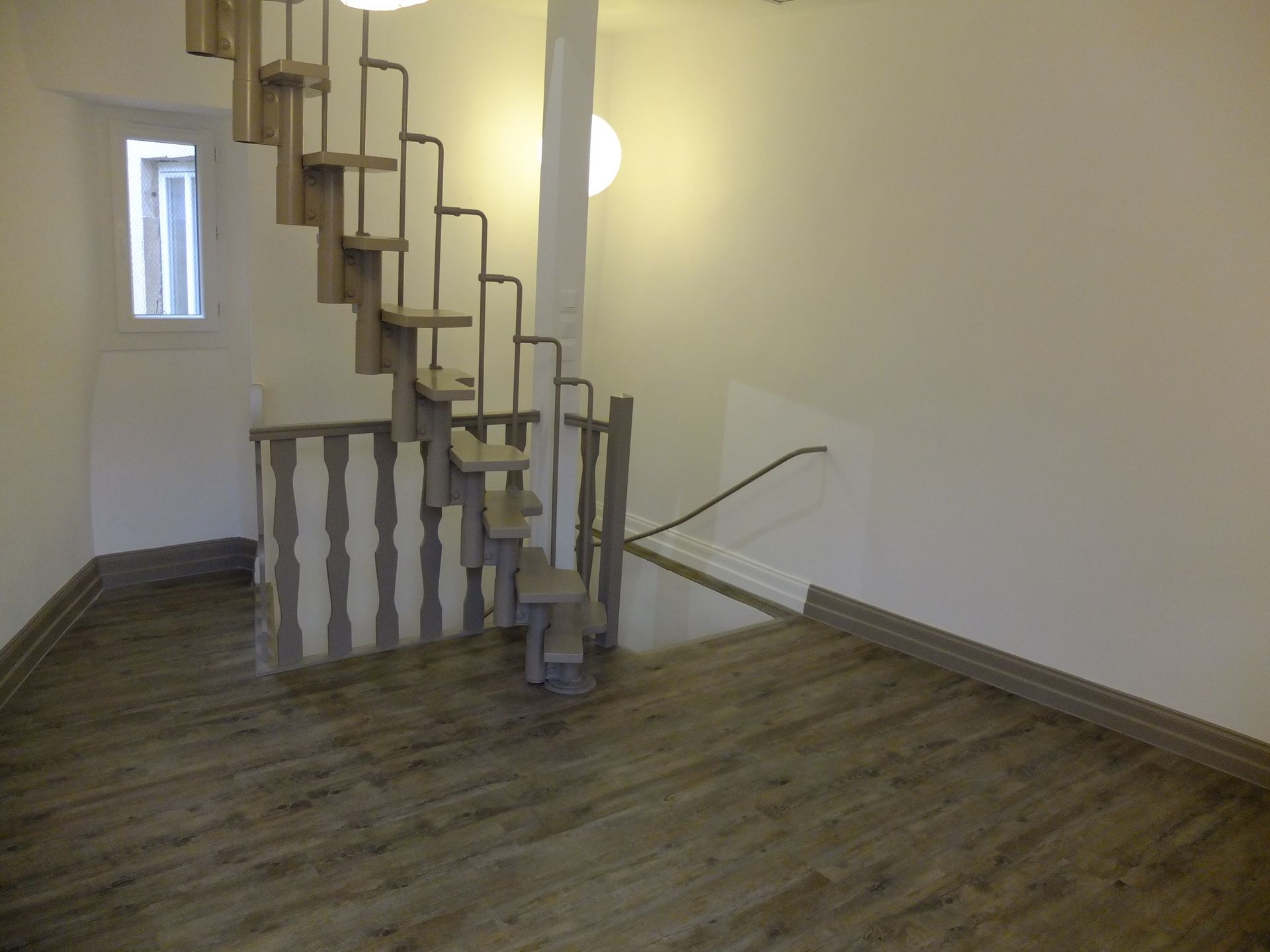 Escalier en bois dans une pièce aux sols clairs et aux murs blancs. Petite fenêtre sur la gauche.