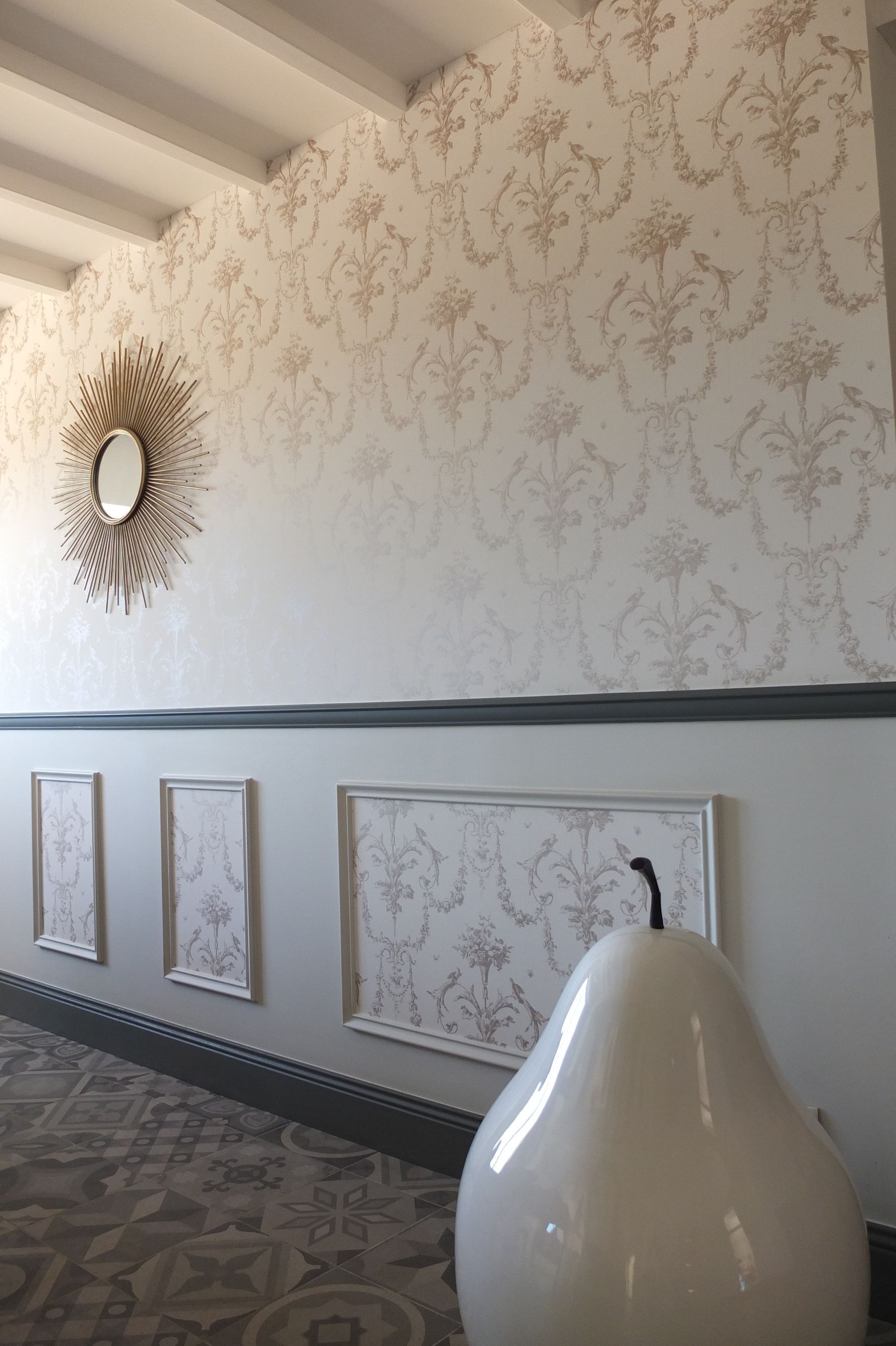 Couloir avec papier peint à motifs, miroir décoratif, panneaux encadrés et sculpture de poire.