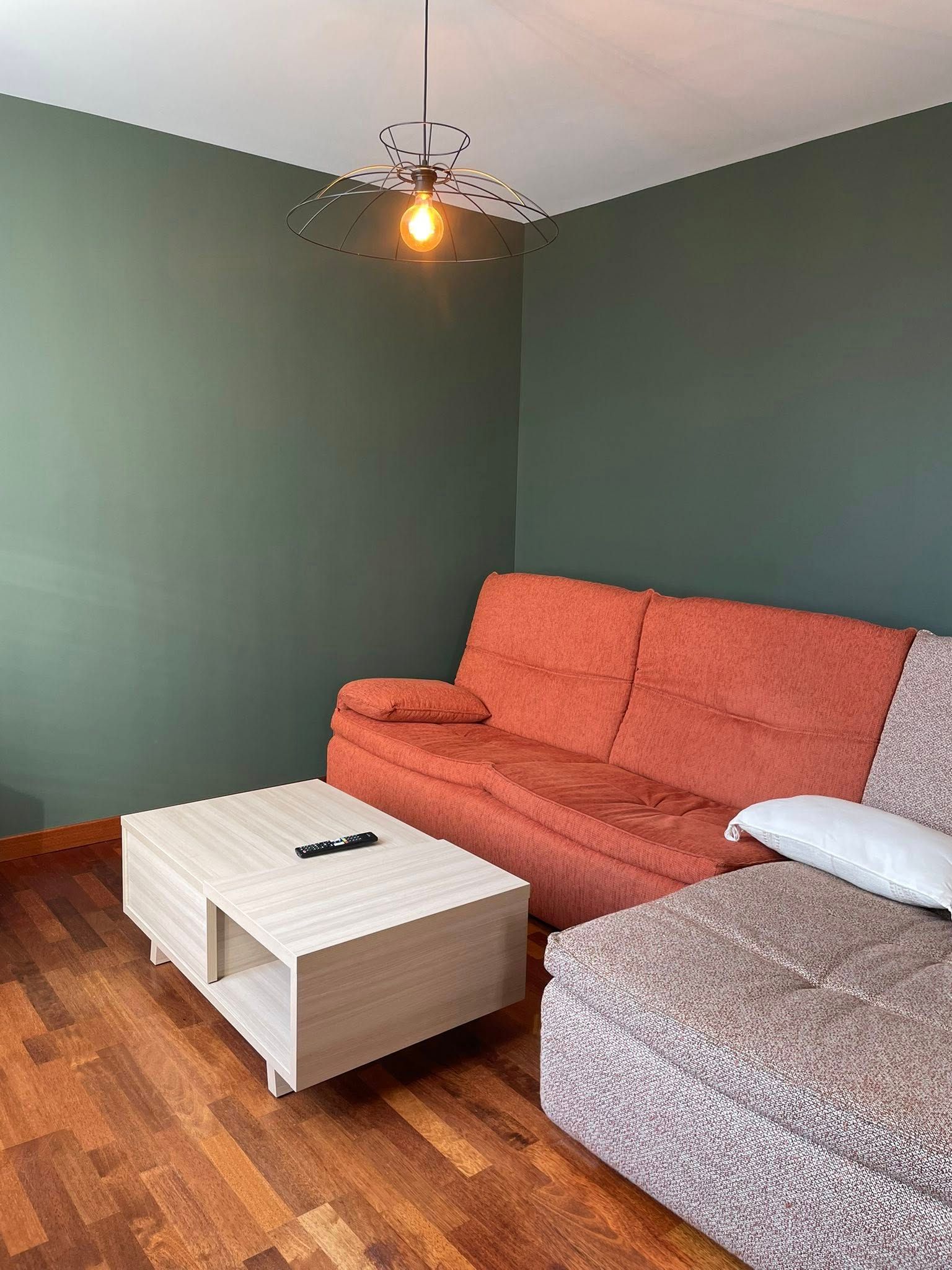 Une table basse moderne en bois clair est placée devant un canapé orange rustique, sur fond de murs vert foncé et de parquet.