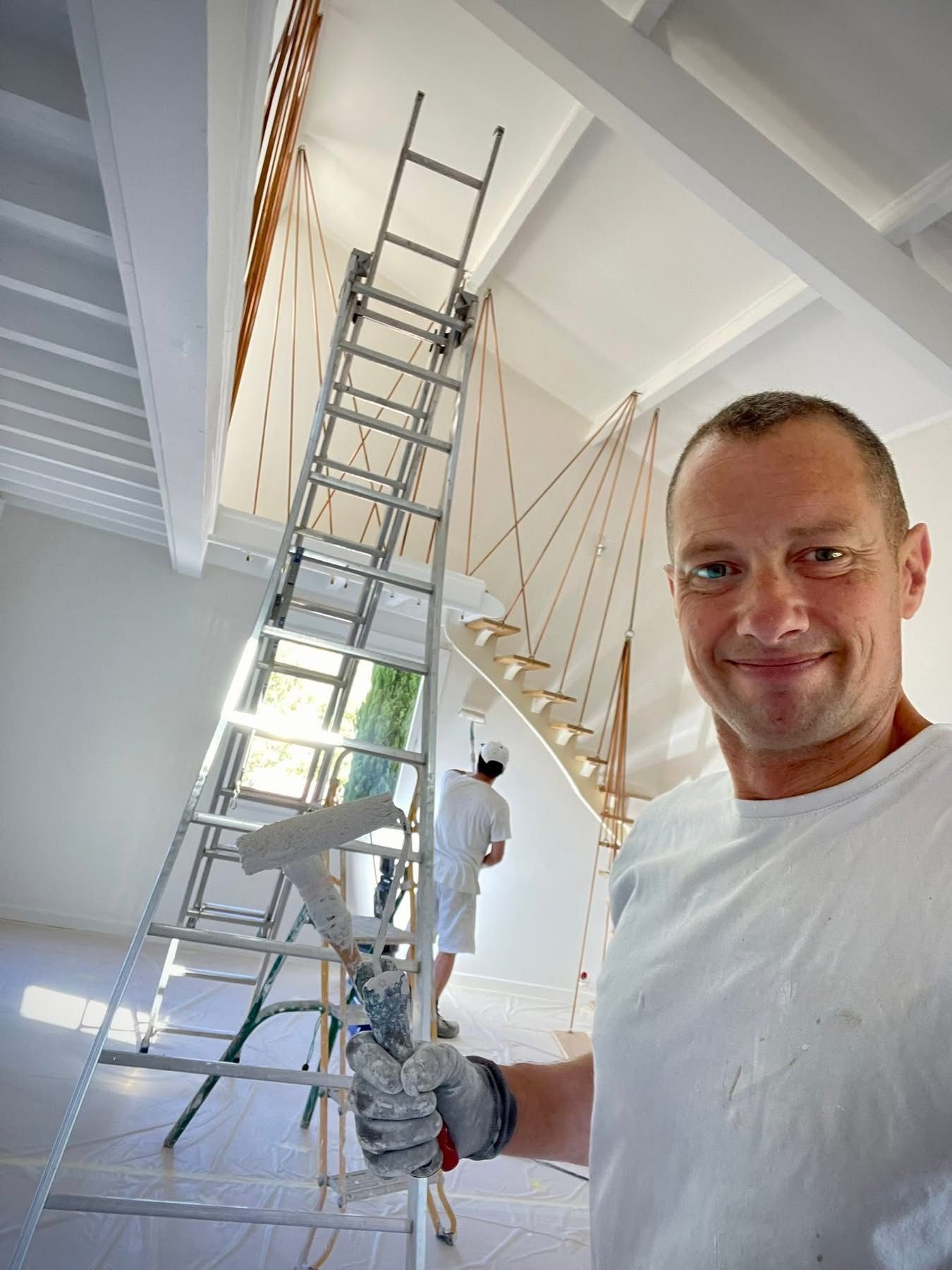 Un homme en chemise blanche, souriant, tient un outil de peinture et se tient près d'une grande échelle.