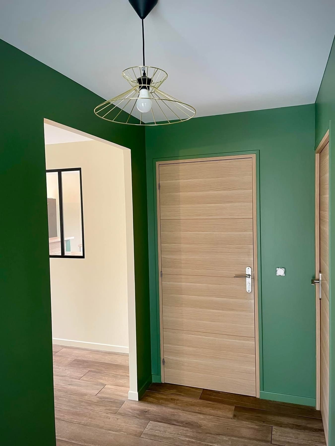 Couloir vert avec une porte en bois clair. Un luminaire décoratif est suspendu au plafond blanc.