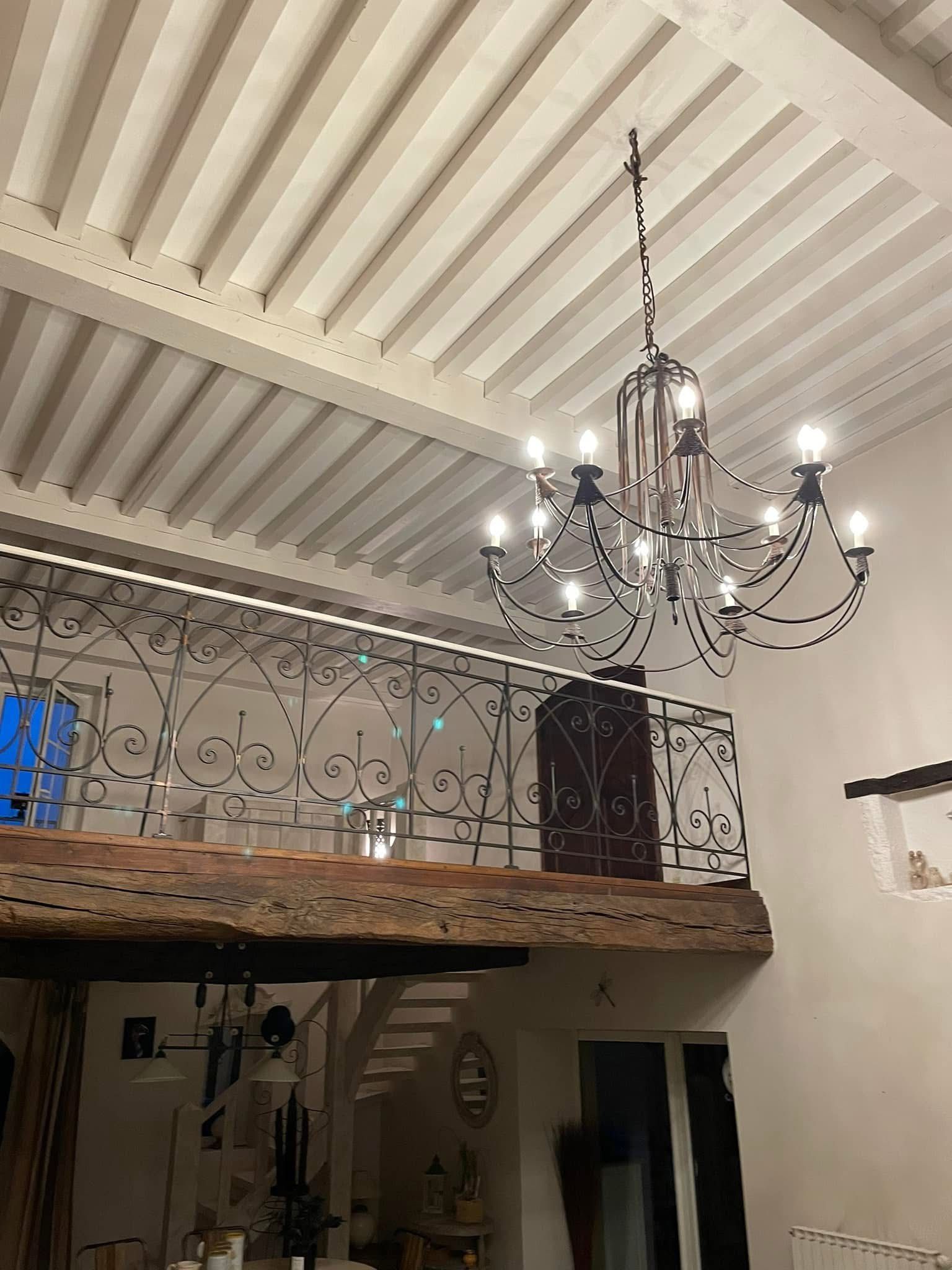 Un lustre est suspendu à un plafond aux poutres blanches. On aperçoit une mezzanine avec une rambarde en fer forgé noir.