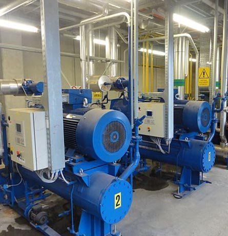 En una sala mecánica se ven enfriadores industriales azules, tuberías y equipos.