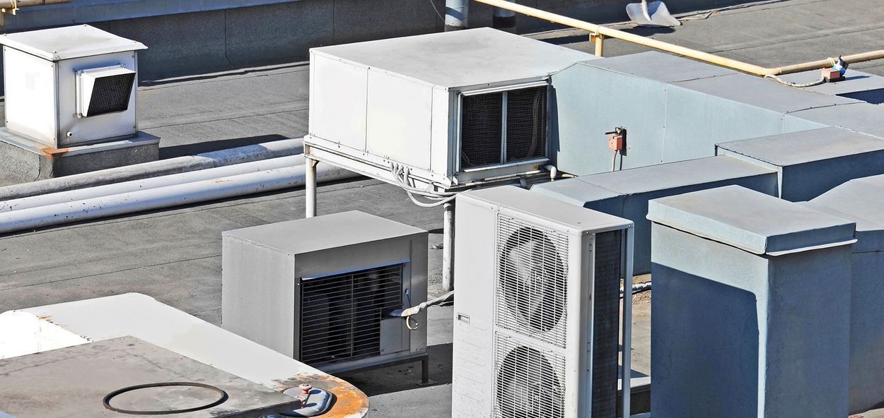 Unidades de aire acondicionado en un tejado. Cajas metálicas grises, ventiladores y tuberías. Luz natural brillante.