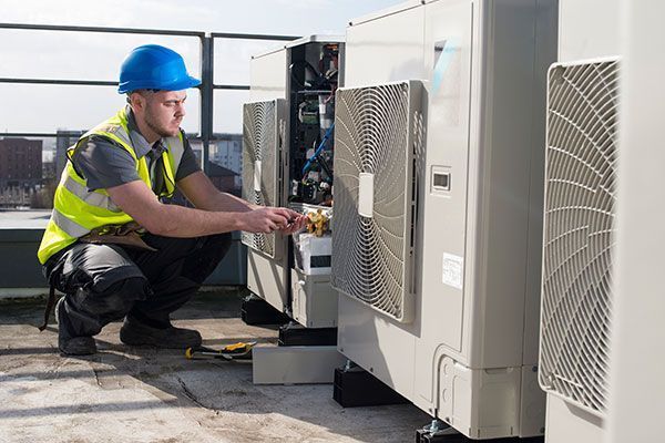 Técnico de HVAC, con casco y chaleco de seguridad, repara unidades de aire acondicionado en azotea.