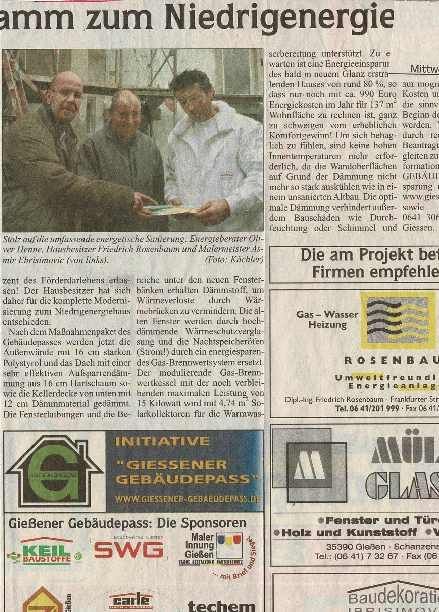 Ein Zeitungsausschnitt zeigt drei Personen, die vor einer Baustelle, umgeben von Werbeplakaten, ein Dokument durchsehen.