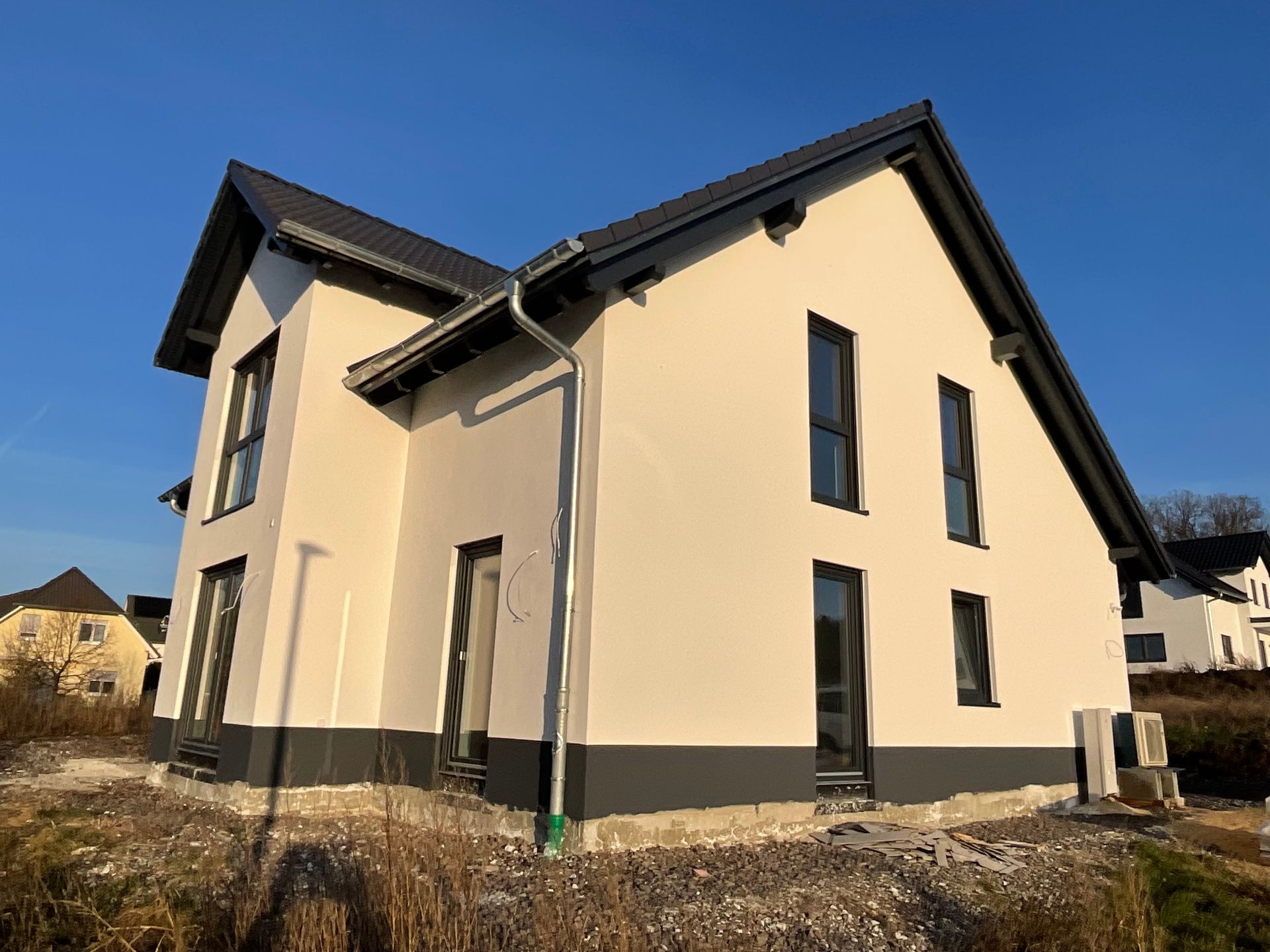 Ein neu errichtetes zweistöckiges Haus mit beigen Wänden, einem dunkelgrauen Sockel und einem dunklen Dach, vor einem klaren blauen Himmel.