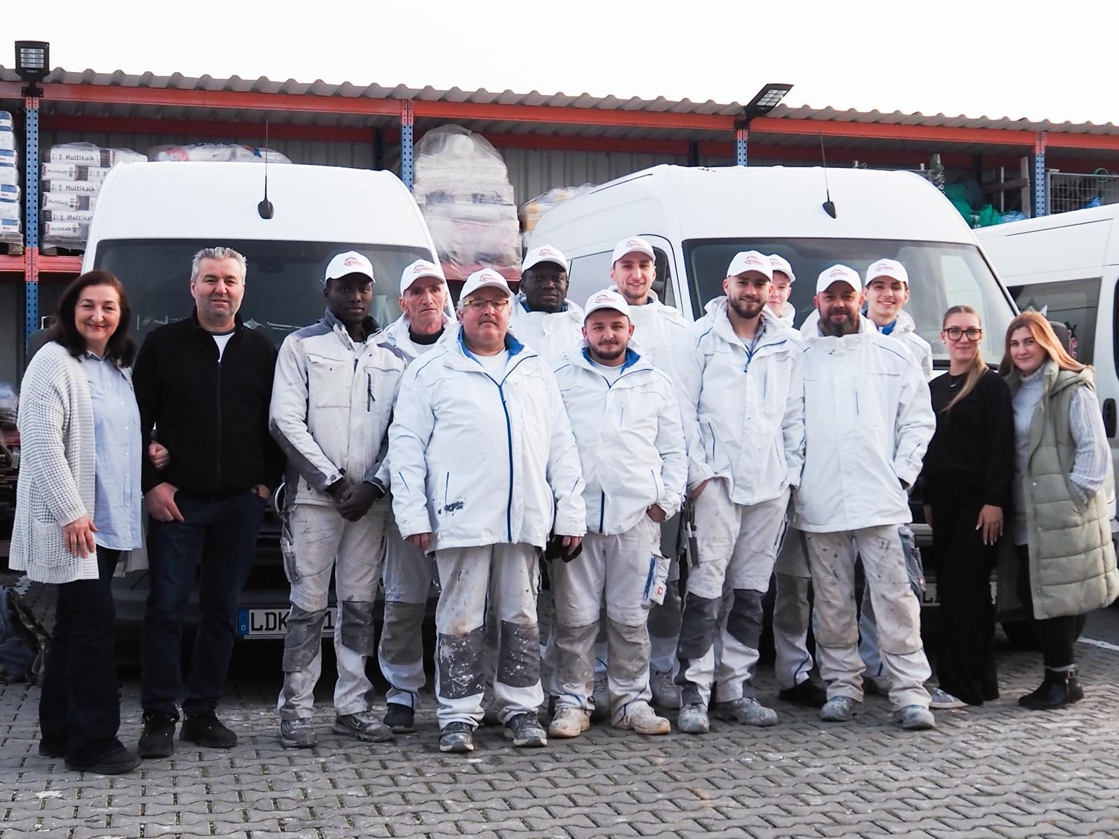 Eine Gruppe von Menschen in weißer Arbeitskleidung steht vor Lieferwagen; einige lächeln.