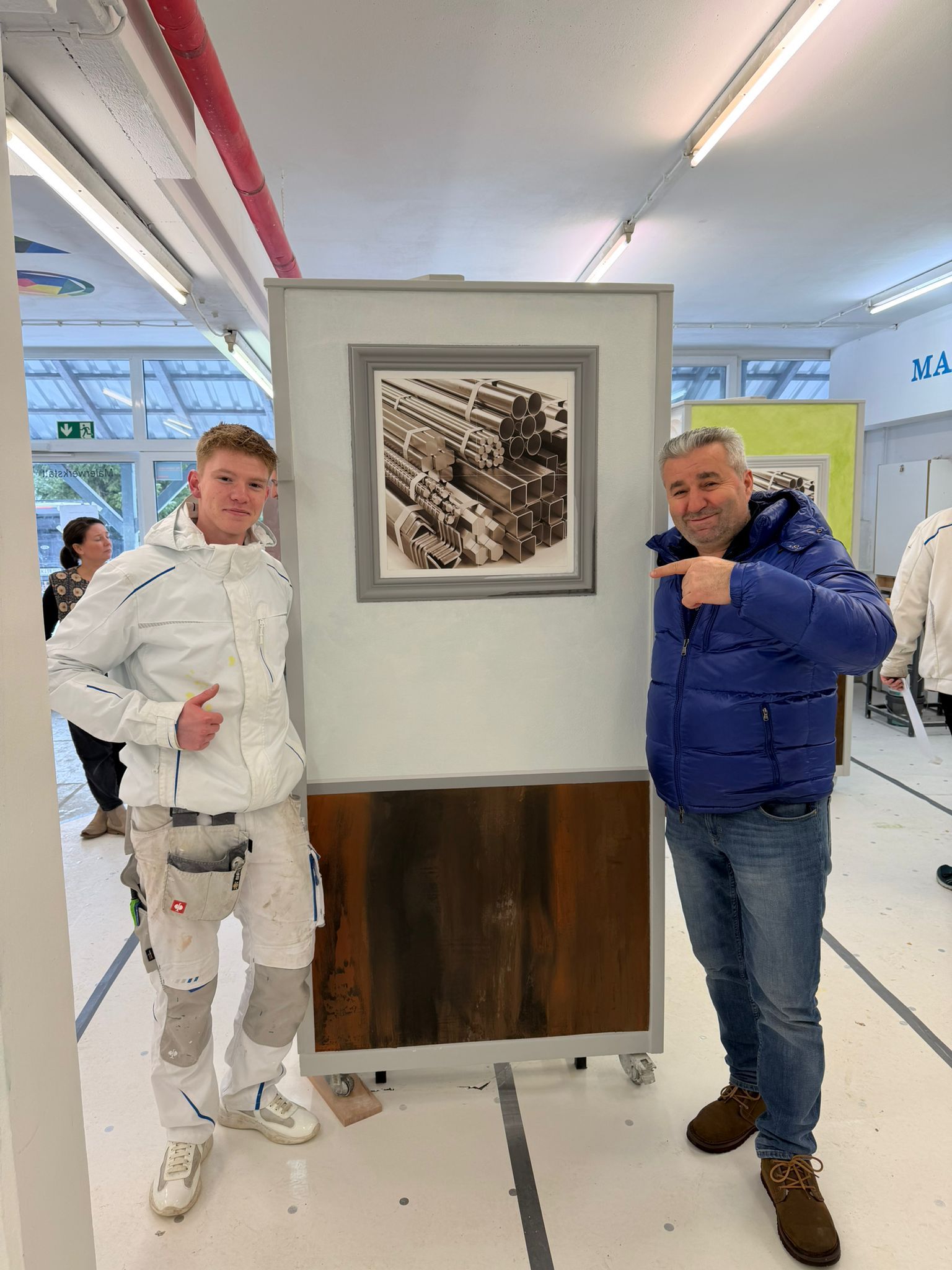 In einer hellen Werkstatt oder einem Atelier stehen zwei Personen jeweils zu beiden Seiten eines ausgestellten Gemäldes.