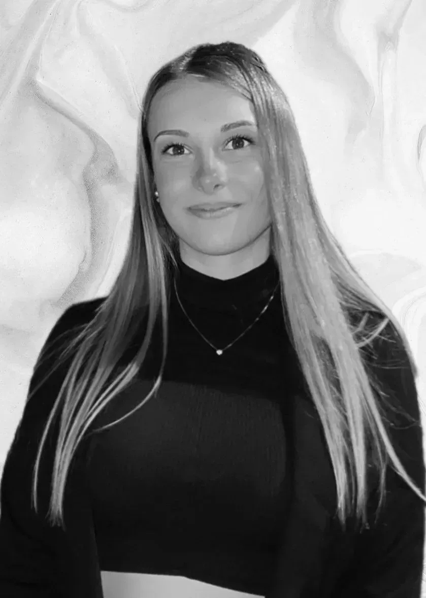 Femme aux longs cheveux blonds, vêtue d'un col roulé et d'un blazer noirs, souriante. Fond blanc marbré.