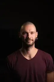 Un homme chauve, barbu et portant des piercings aux oreilles, sourit, vêtu d'une chemise bordeaux à col en V, sur fond noir.