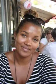 Femme avec des lunettes de soleil sur la tête, souriante dans une chemise à rayures, assise à une table en extérieur.