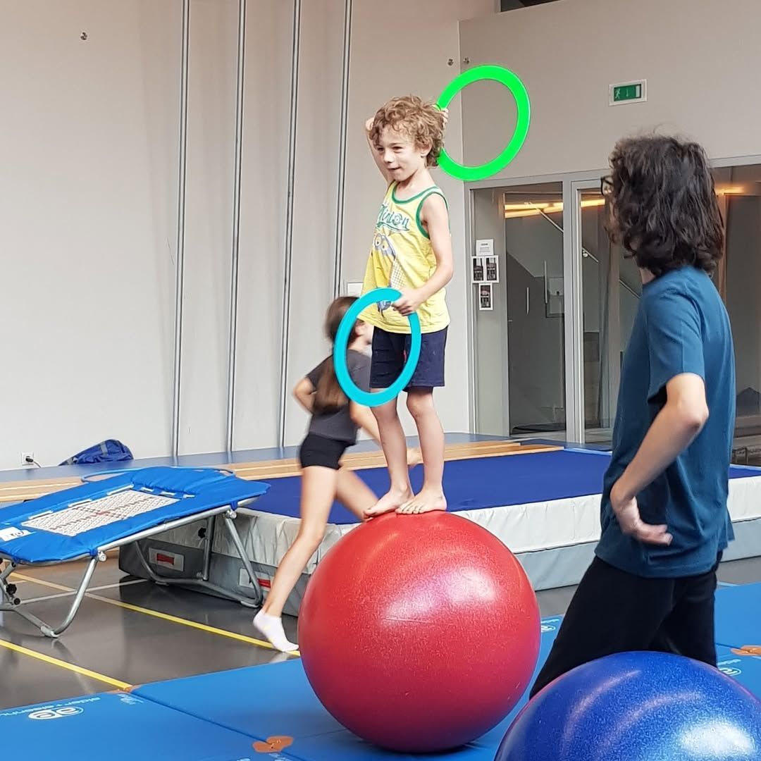 Un garçon en équilibre sur un ballon, tenant des anneaux, soutenu par une fille. Une femme les observe dans une salle de sport.