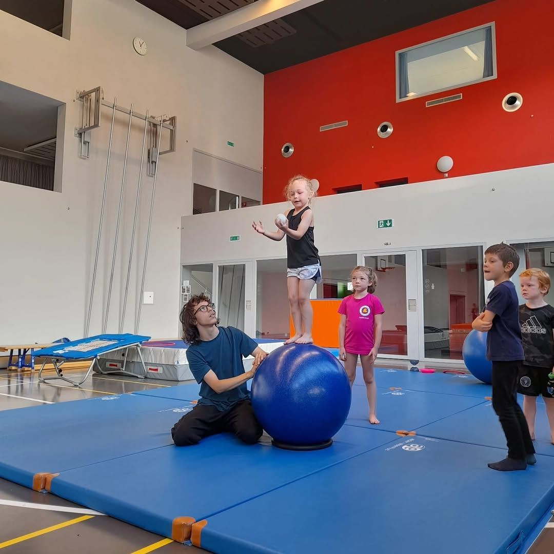Des enfants en équilibre sur des ballons d'exercice dans une salle de sport, avec l'aide d'un moniteur.