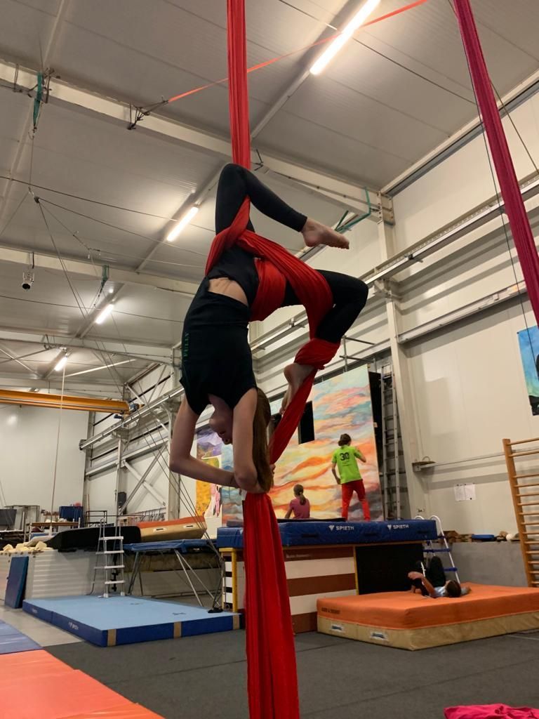 Une personne pratiquant un acrobatie aérienne sur tissu rouge, sous le regard de spectateurs prenant des photos dans une salle de sport.