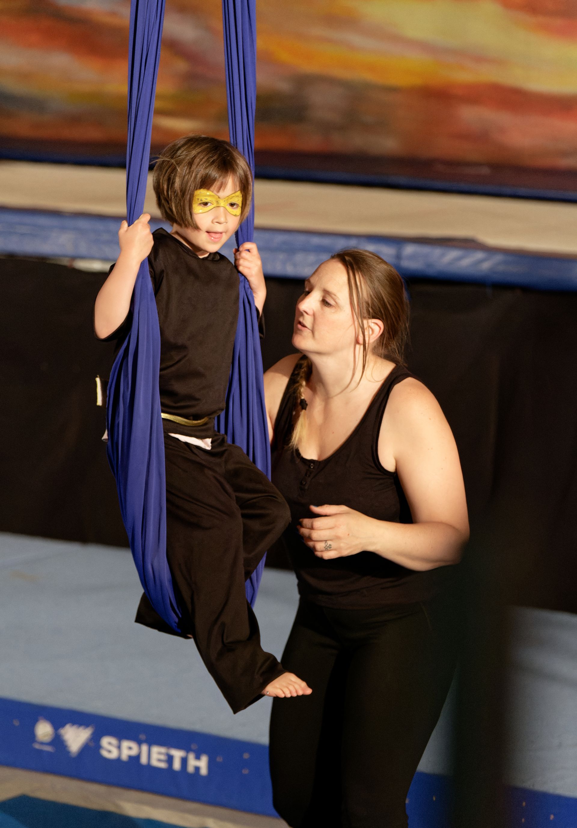 Salle d'entraînement de cirque : une acrobate aérienne dans les airs, d'autres spectateurs au sol ; tapis, cordes et une grande pile de tapis.