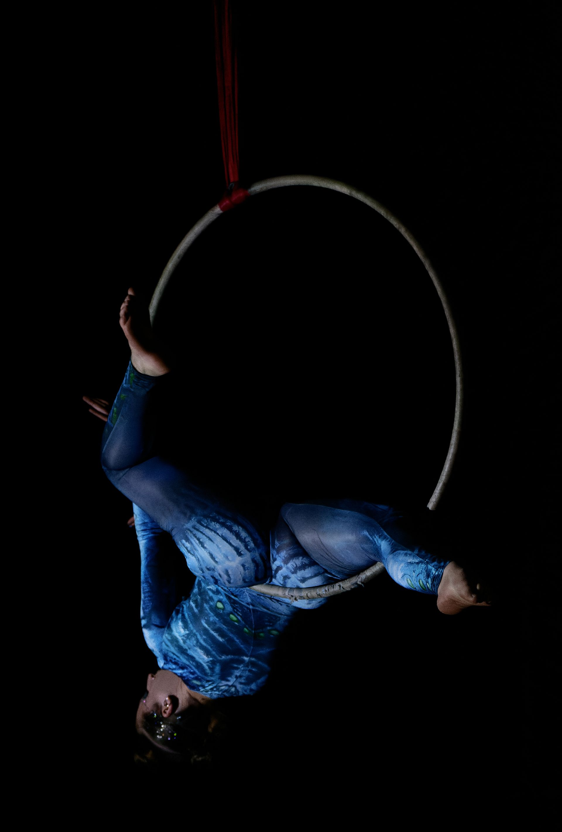 Une personne pratiquant un acrobatie aérienne sur tissu rouge, sous le regard de spectateurs prenant des photos dans une salle de sport.