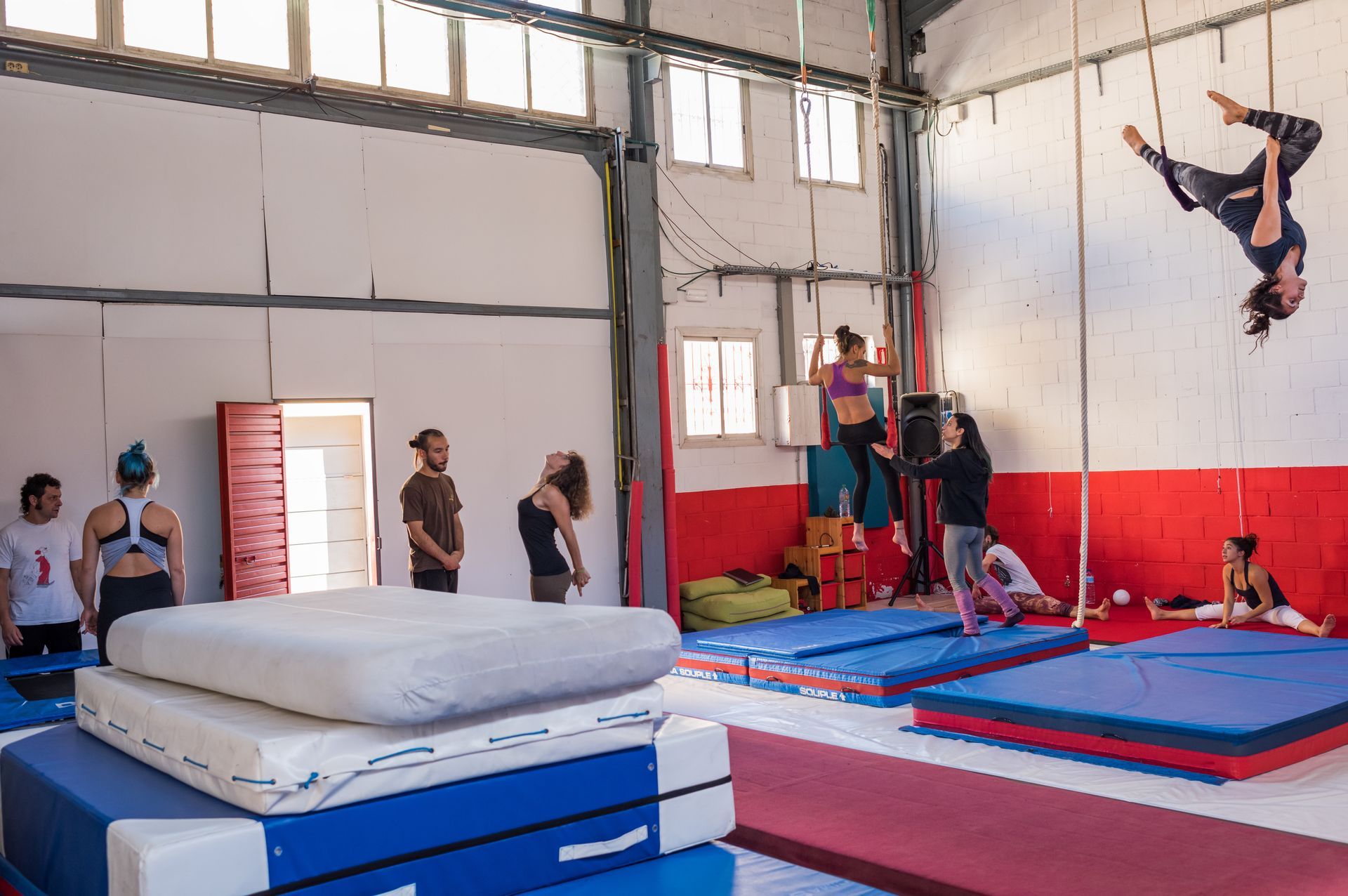 Salle d'entraînement de cirque : une acrobate aérienne dans les airs, d'autres spectateurs au sol ; tapis, cordes et une grande pile de tapis.