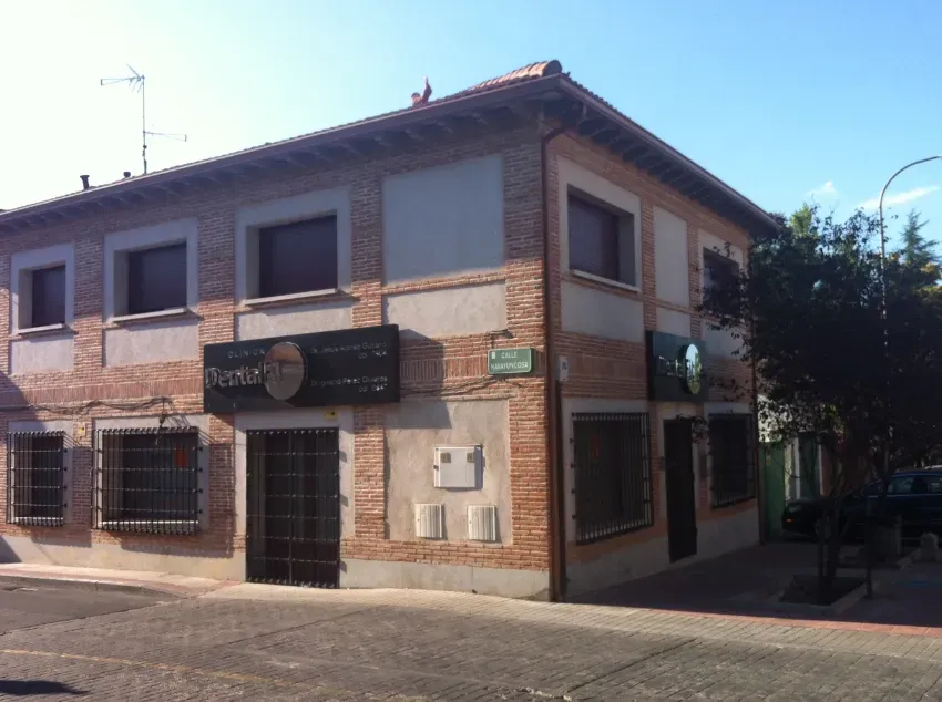 Edificio esquinero de ladrillo y dos plantas, con las ventanas cerradas con contraventanas y un letrero oscuro en el local, en una calle soleada.