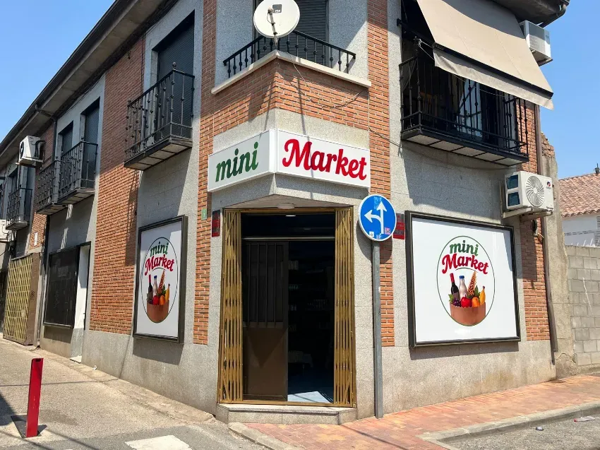 Fachada de una pequeña tienda de barrio, con letreros rojos y verdes, en un edificio de ladrillo situado en una esquina