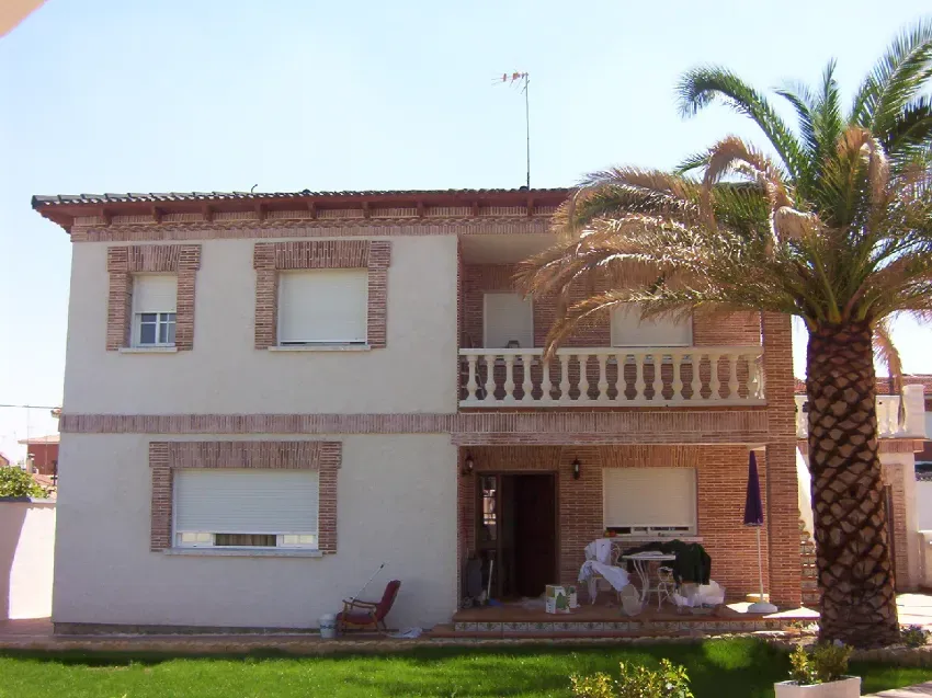 Casa blanca de dos plantas con detalles de ladrillo, balcón y palmeras delante, bajo un cielo despejado