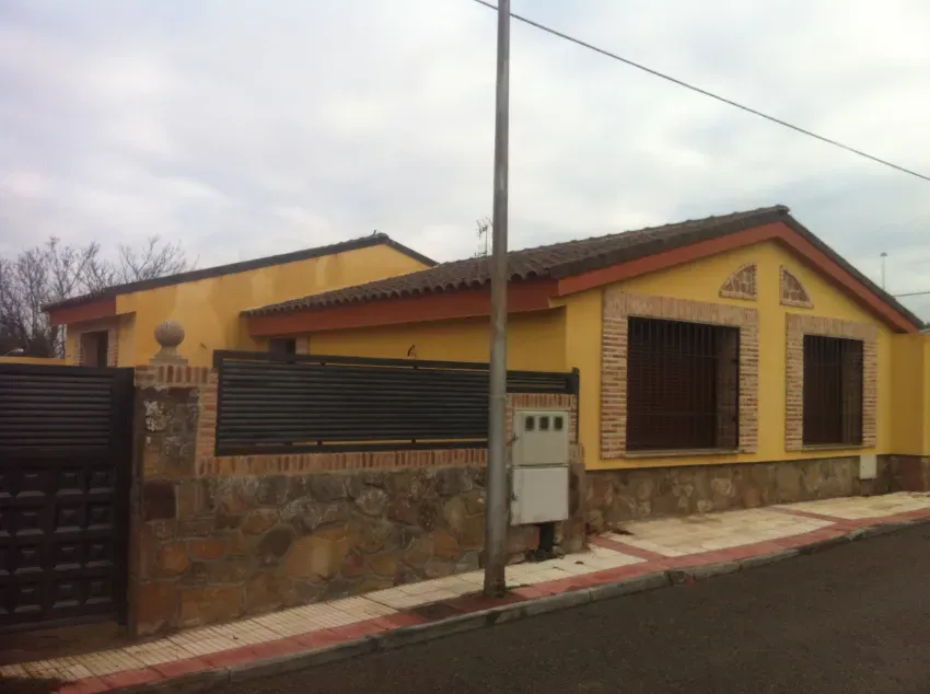Casa amarilla con tejado rojo y ventanas enrejadas, detrás de un muro de piedra y una verja metálica