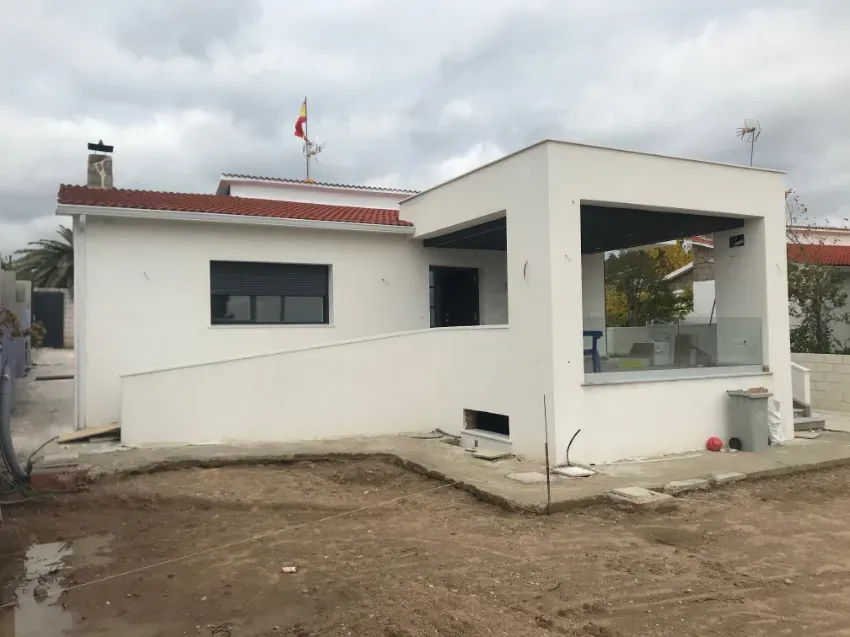 Pequeña casa blanca con techo rojo en construcción, con un porche delantero cubierto y un patio de tierra sin vegetación.