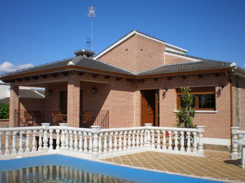 Casa de ladrillo con tejado de tejas y balaustrada blanca junto a una piscina bajo un cielo azul despejado
