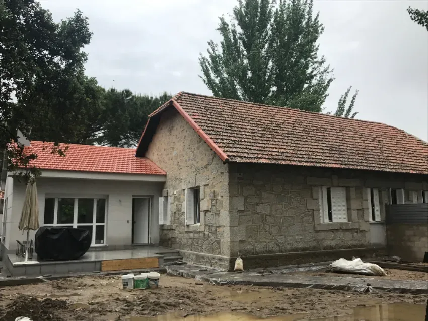 Casa de piedra en renovación, con tejados de tejas rojas, ventanas blancas y escombros de obra delante