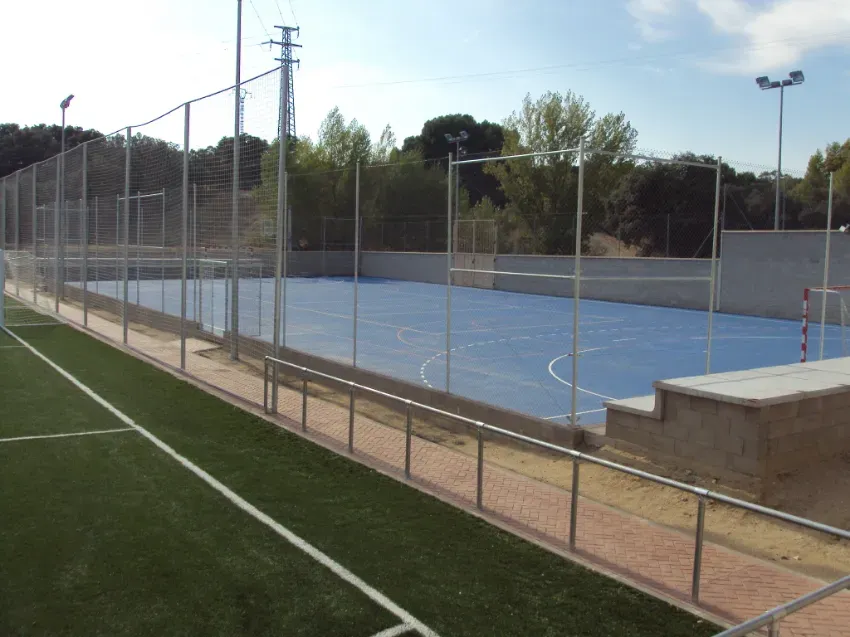 Cancha deportiva exterior azul vacía junto a un campo de fútbol verde, rodeada por una valla metálica.
