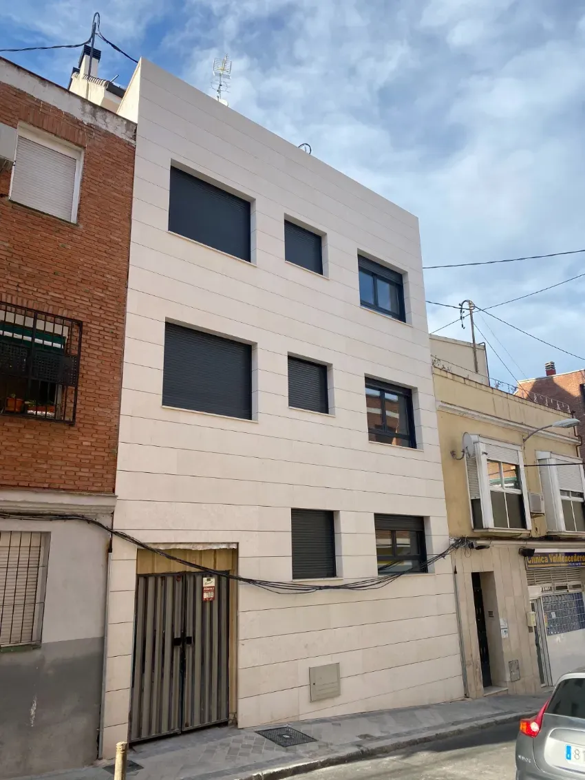 Edificio de apartamentos estrecho de tres plantas, con fachada blanca y ventanas oscuras, en una calle residencial