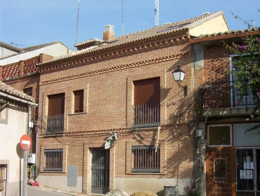 Fachada de una casa adosada de ladrillo, con ventanas enrejadas, un balcón y una farola en una calle soleada