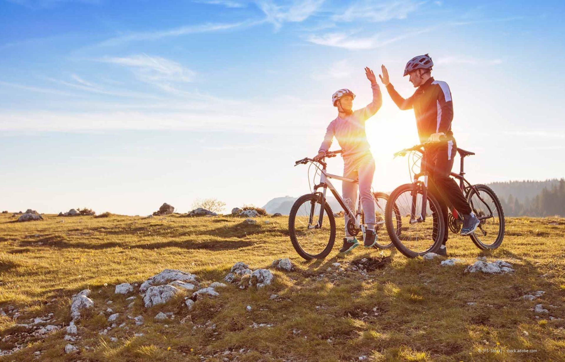 Zwei Radfahrer geben sich bei Sonnenuntergang auf einem grasbewachsenen Hügel neben ihren Fahrrädern ein High-Five.