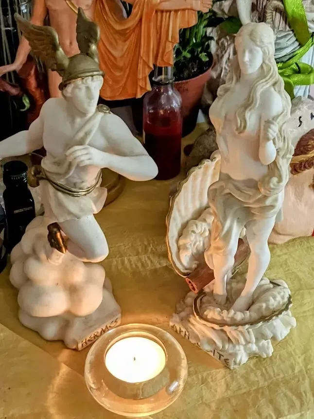 Estatuillas de los dioses Hermes y Afrodita frente a una vela de t&eacute;. 