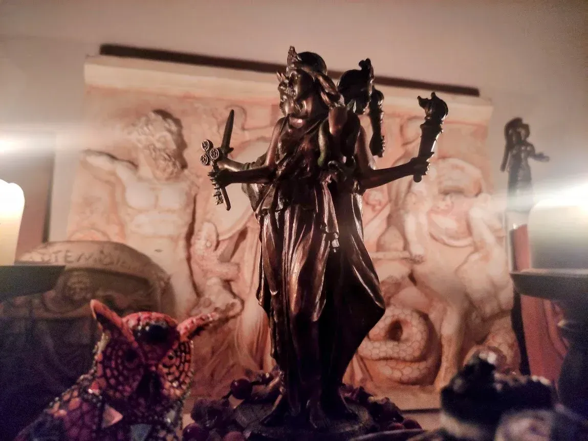 Estatua oscura con cuernos y brazos extendidos frente a figuras de piedra talladas y luz de velas.