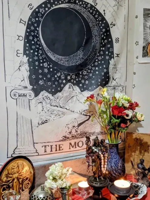 Un tapiz con motivos de luna y estrellas se encuentra detrás de un pequeño altar con flores, velas y jarrones decorativos.