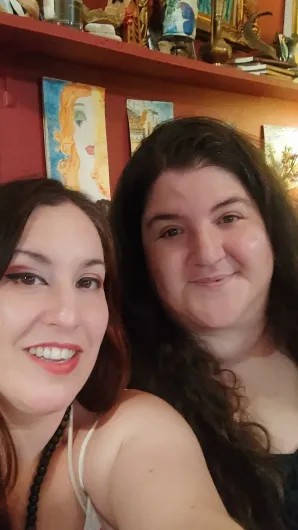 Dos mujeres sonriendo para una selfie frente a una pared colorida con estantes y elementos decorativos.