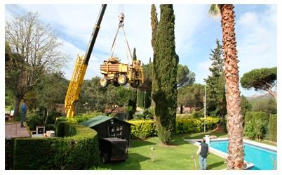 Grúa amarilla levantando un gran vehículo amarillo sobre un patio trasero con piscina.