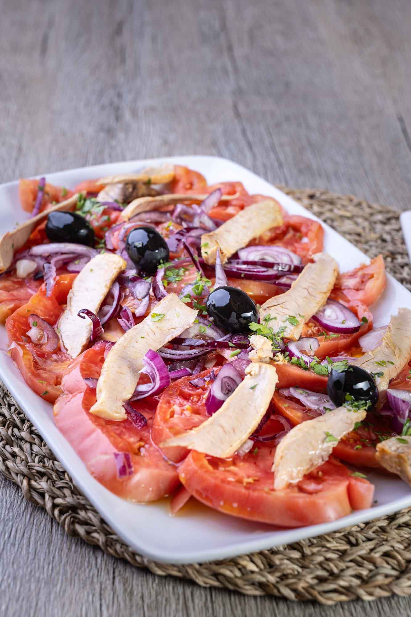 Un plato blanco cubierto con tomates, cebollas, aceitunas y atún.