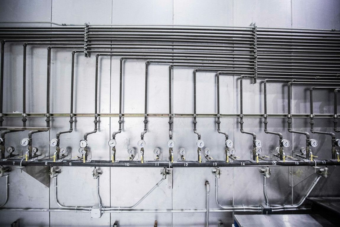 Una hilera de tuberías industriales de acero inoxidable, válvulas y manómetros montados contra una pared de azulejos grises.
