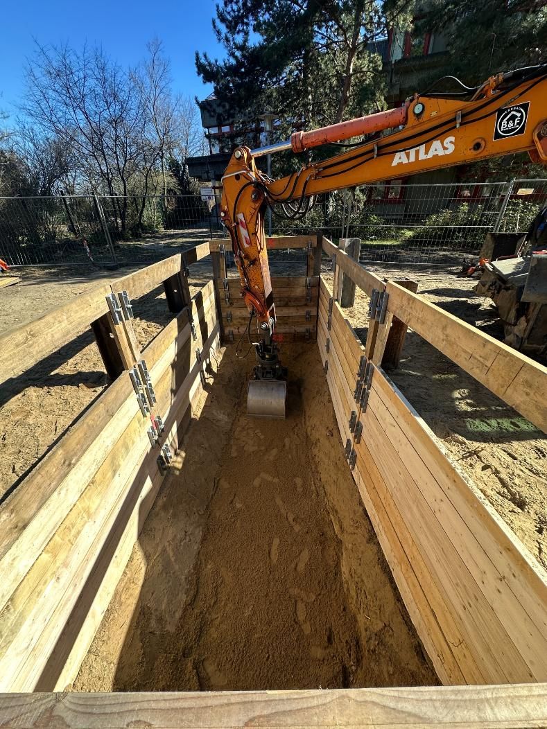Ein Bagger gräbt in einem langen, mit Holz ausgekleideten Graben. Sonnige Außenaufnahme.