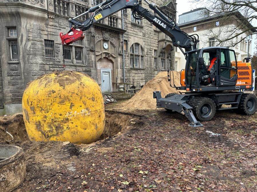 Ein Bagger hebt einen großen, gelben, kugelförmigen Gegenstand aus einem Loch im Boden in der Nähe eines Gebäudes.