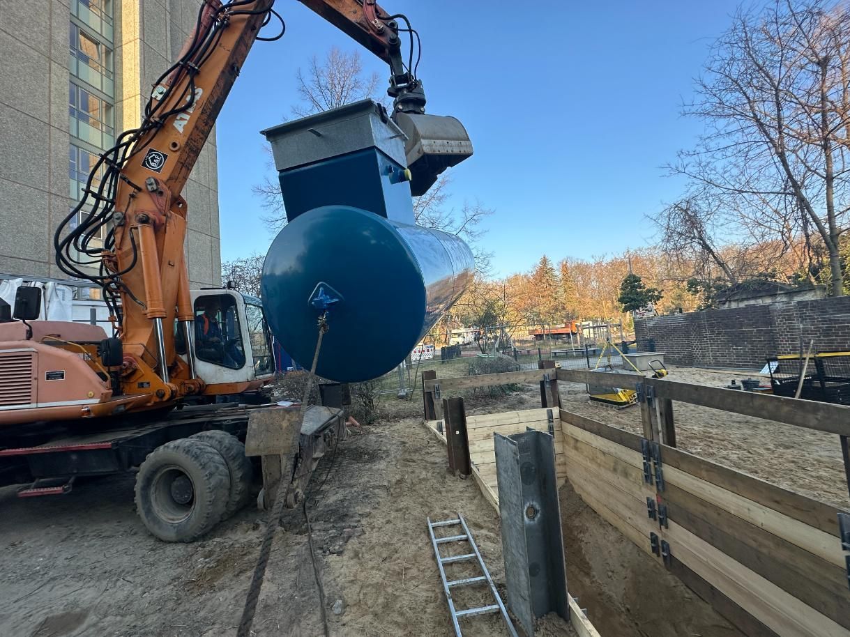 Ein Bagger hebt einen großen blauen Tank an. Baustelle mit Bäumen und Gebäuden im Hintergrund.