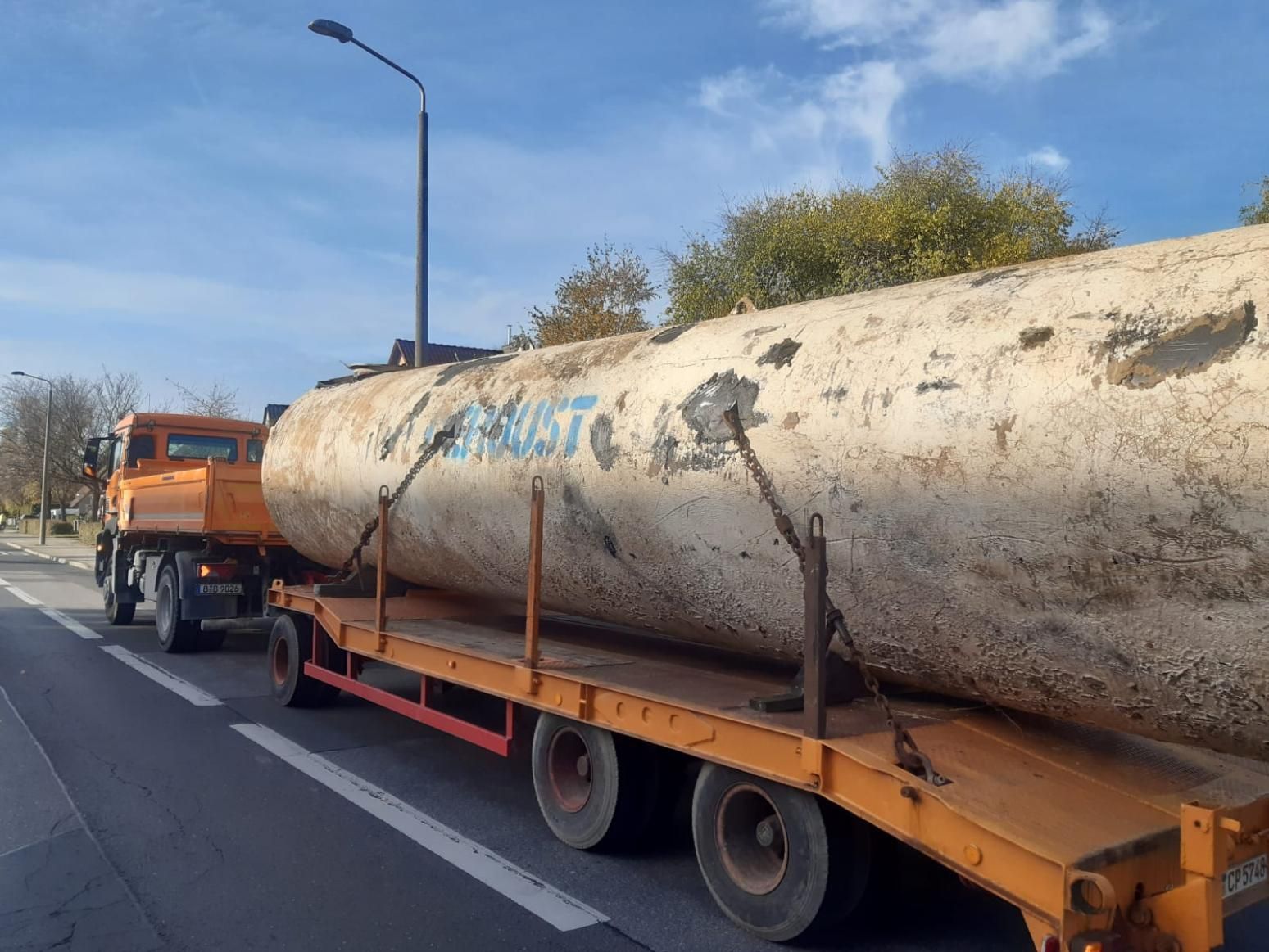 Ein Pritschenwagen transportiert an einem sonnigen Tag einen großen, verwitterten zylindrischen Gegenstand über eine Straße.