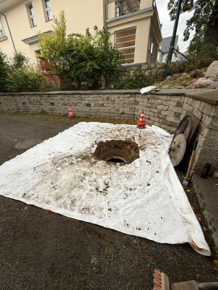 Ein offener, mit einer weißen Plane abgedeckter Kanaldeckel ist von Schutt umgeben. 