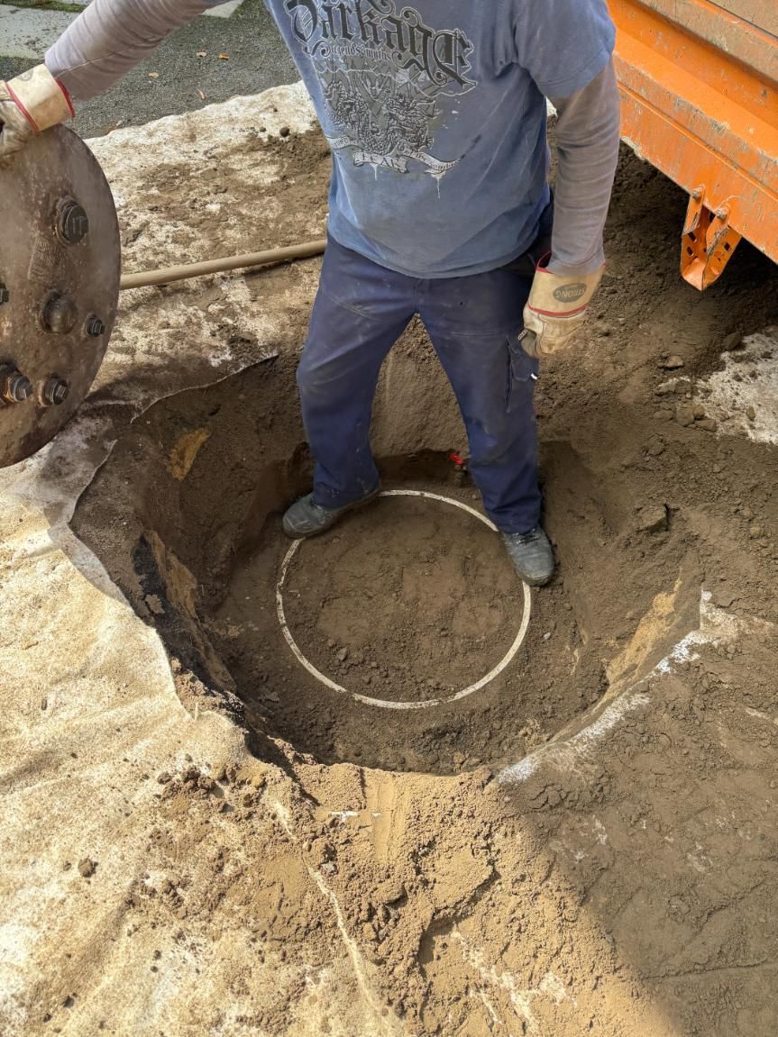 Eine Person sitzt in einem Loch und hält einen Kanaldeckel neben einem weißen Kreis. 