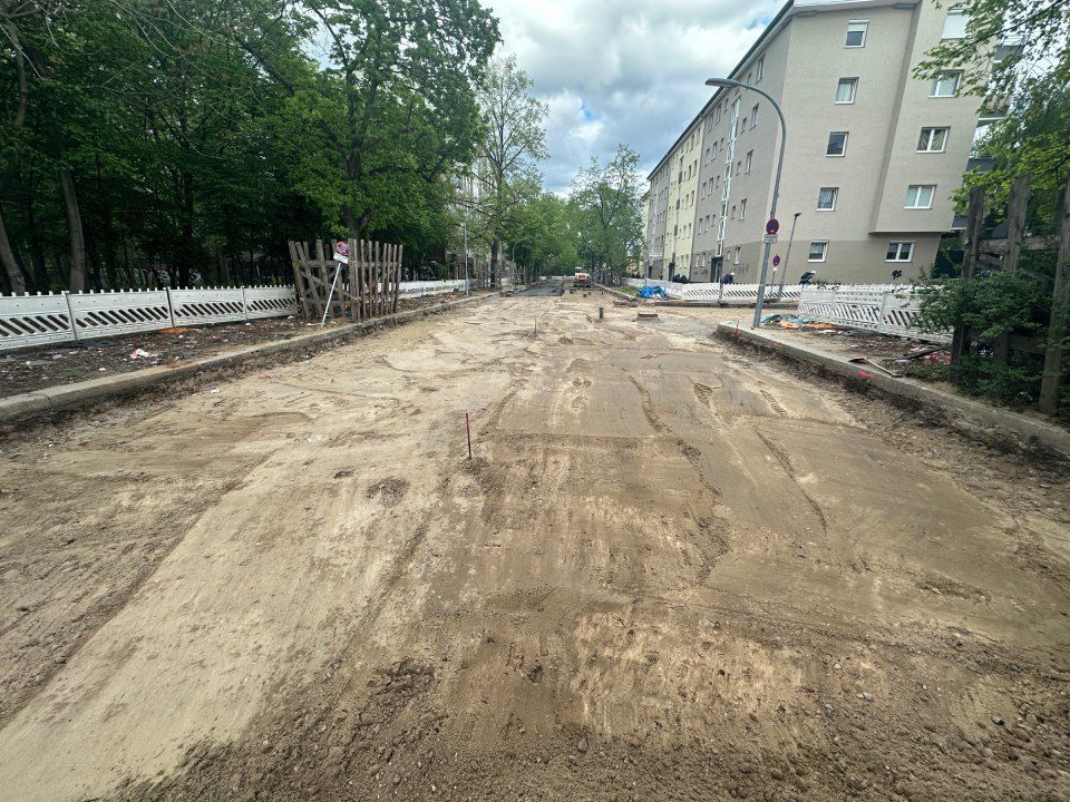 Straßenbaustelle mit unbefestigter Straße, Gebäuden, Bäumen und bewölktem Himmel.