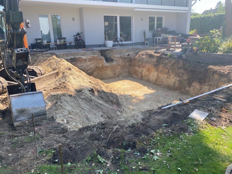 Aushebung einer rechteckigen Grube in einem Hinterhof für einen Pool, links ein Bagger und im Hintergrund ein Haus.