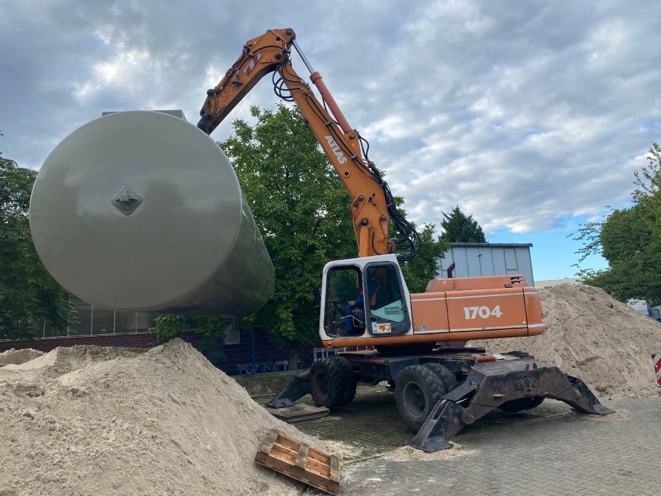 Ein orangefarbener Bagger hebt einen großen, zylindrischen, grauen Tank auf einer Baustelle an.