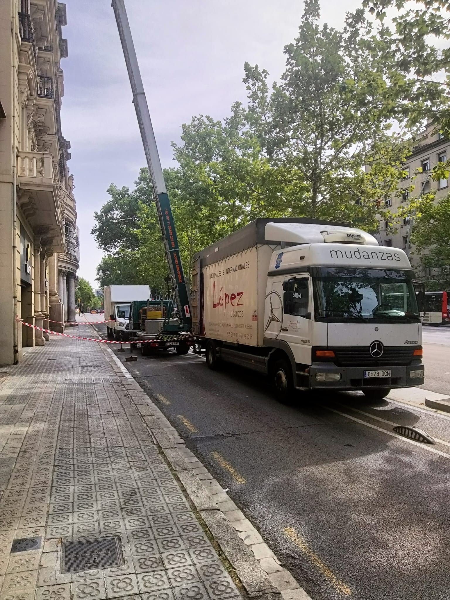 Camión con plataforma elevadora en una calle de la ciudad, cargando o descargando. Edificio alto a la izquierda, árboles y autobús al fondo.